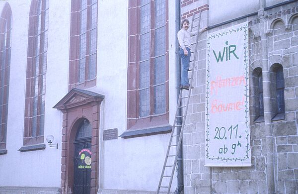 An lächelnder man steht auf einer an eine Kirchenfassade gelehnten Leiter. Anscheinend hat er gerade ein bunt beschriebenes Bettlaken an der Fassade angebracht. Auf diesem Transparent steht: "Wir pflanzen bäume, 20.11., ab 9 Uhr". 