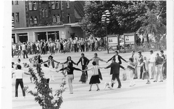 Schwarz-Weiß-Foto:Eine Gruppe von Menschen tanzt händehaltend auf einem Platz im Kreis. Im Hintergrund hat sich eine Menschenmenge zum Zuschauen gebildet.