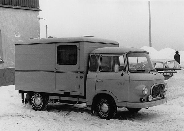 Schwarz-Weiß-Foto eines Kastenwagens bzw. Transporter, der vor einem Gebäude auf einem verschneiten Parkplatz steht.