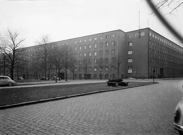 Blick über einen winterlichen Platz auf ein grau verputztes, sechsgeschossiges Gebäude Platz