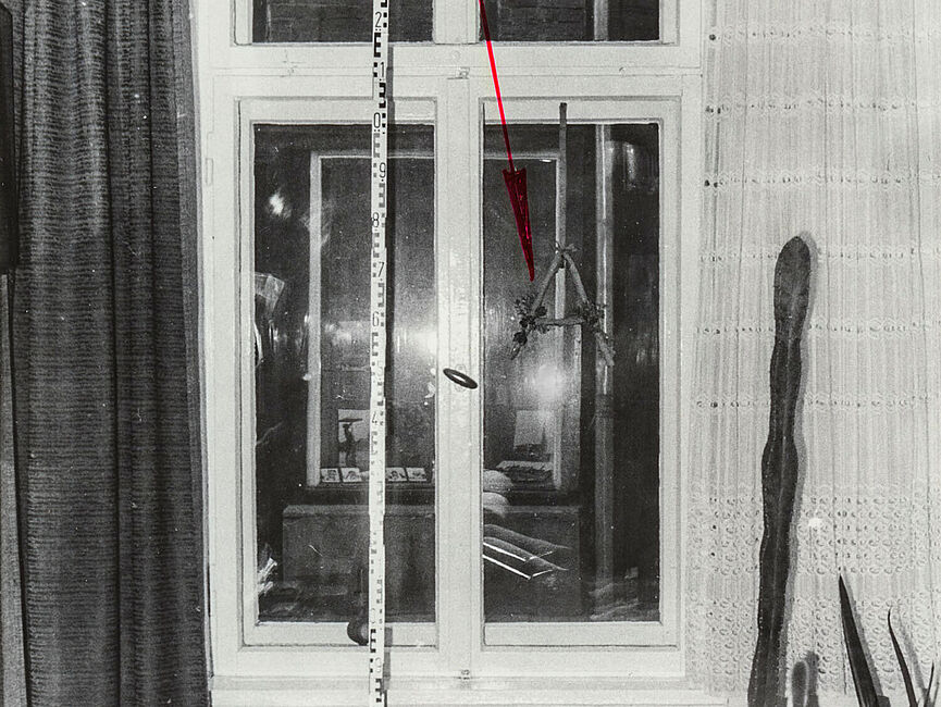 Schwarz-Weiß-Foto eines Fensters in einem Wohnzimmer. Ein eingezeichneter roter Pfeil zeigt auf ein gebasteltes "A", das am Fensterrahmen hängt. Daneben ist ein Maßband.