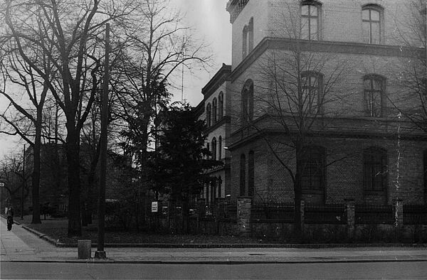 Das Bild ist über eine Straße fotografiert. Es zeigt ein Fabrikgebäude aus der Gründerzeit. Die Wandflächen bestehen aus beigen Ziegeln, Gesimse aus rot-braunen Ziegelelementen. Davor stehen Bäume ohne Laub.