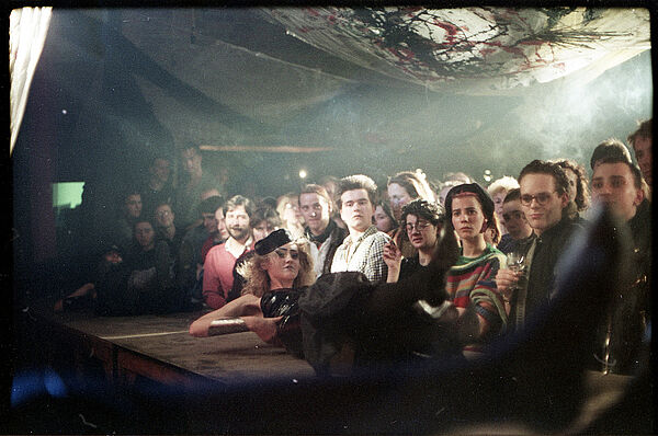 Modeperformance der Künstlergruppe „chic, charmant & dauerhaft“, Berlin 1987; Foto: Hartmut Beil Auf einer Bühne in einem verrauchten Raum liegt ein Model im auffälligen Kostüm. Vor der Bühne stehen dicht gedrängt Zuschauer, manche Blicken zum Fotografen.