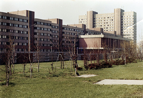 BV Berlin ab 1985, Straße der Befreiung 60 (heute Alt-Friedrichsfelde 60) Blick über eine Grünfläche mit frisch gepflanzten Bäumen auf das aus einem Gebäuderiegel und -Turm bestehende, in Plattenbauweise errichtete BV-Gebäude