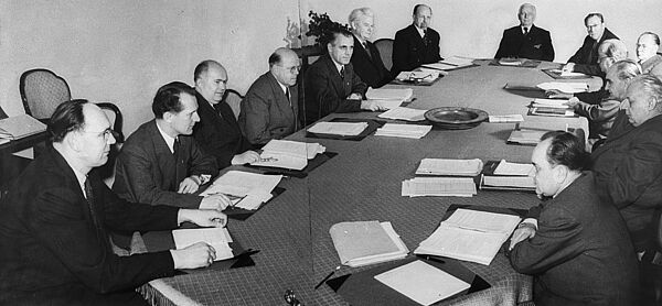 Sitzung des Politbüros der SED, April 1955; Quelle: BArch, Bild 183-30033-0002, Fotograf: Walter Heilig Schwarz-weiß Foto, das aus zwei Einzelfotos zu einem Panorama zusammengefügt wurde: Die ausschließlich männlichen Mitglieder des Politbüros sitzen um einen langgestreckten Konferenztisch herum, jeweils vor ihnen liegen ihre Arbeitspapiere auf dem Tisch.