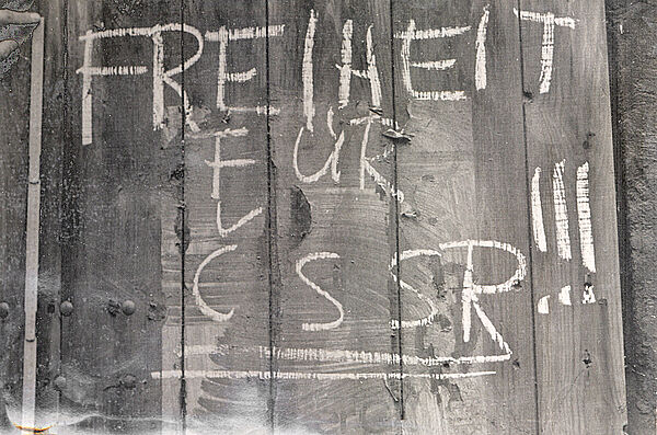 Schwarzweißbild: auf einer Oberfläche aus Holzbrettern sind weiße Kreidelettern zu sehen: "Freiheit für CSSR". Links daneben ist ein Zollstock angelegt, man sieht noch den Teil einer hand oben am Zollstock..