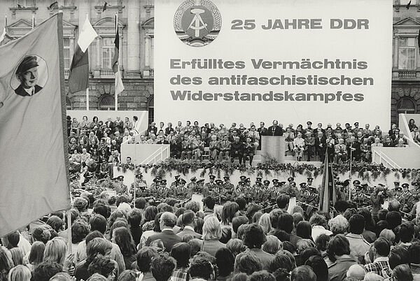 Blick auf das mit Blumen geschmückte Podium einer Kundgebung, auf dem zahlreiche Gäste sitzen. Ein Redner steht am Pult. Das Podium wird von einem Banner überragt. Es trägt neben dem Wappen der DDR den Text: "25 Jahre DDR. Erfülltes Vermächtnis des antifaschistischen Widerstandskampfes. Vor dem Podium sitzt eine Militärkapelle, davor drängen sich die Zuschauer. Im Vordergrund ist auf einer Fahne ein Portrait Ernst Thälmanns zu erkennen.