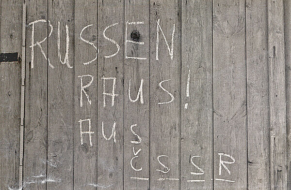 Schwarzweißbild: auf einer Oberfläche aus Holzbrettern sind weiße Kreidelettern zu sehen: "Russen raus aus CSSR". Links daneben ist ein Zollstock angelegt.