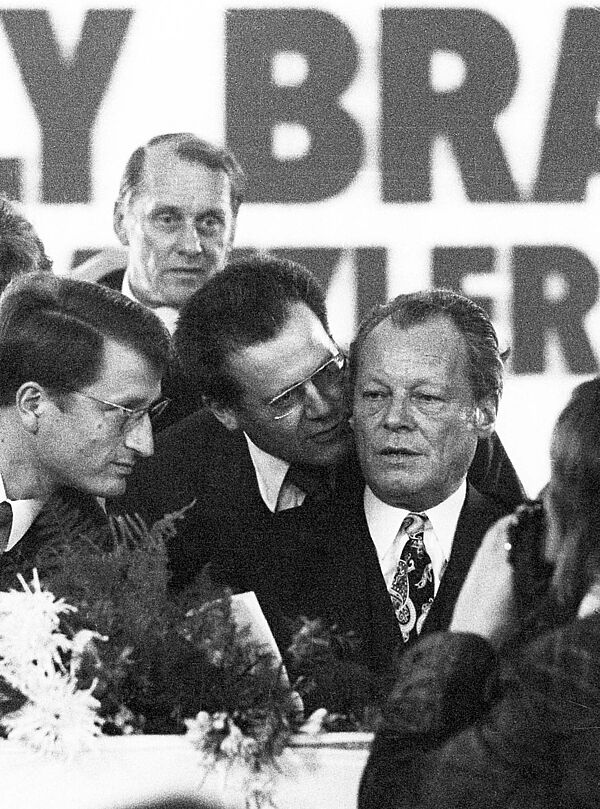 Günter Guillaume (Mitte), Stasi-Spion und persönlicher Referent des Bundeskanzlers Willy Brandt (rechts), auf dem SPD-Parteitag in Dortmund, 12. Oktober 1972 Nahaufnahme Willy Brandts auf einem Podium. Günter Guillaume flüstert dem aufmerksam zuhörenden Brandt etwas in sein rechtes Ohr Ohr.