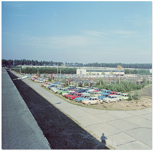 Blick von einem Lagerhallendach über eine weite Fläche voller KFZ mit unterschiedlichen Farben. Im Hintergrund Lagergebäude.