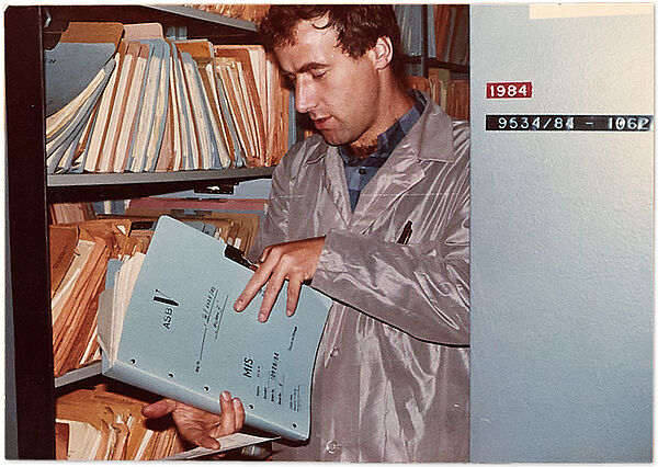 Ein Stasi-Mitarbeiter mit archivierter Akte vor einem Magazinschrank des Zentralarchivs, zwischen 1984 und 1989; Quelle: BArch, MfS, Abt. XII, Nr. 8408; Tafel 2 (Ausschnitt) Foto eines Mannes, der eine aufgeschlagene Akte in der Hand hält. Im Hintergrund ist ein Schrank voller Akten zu sehen.