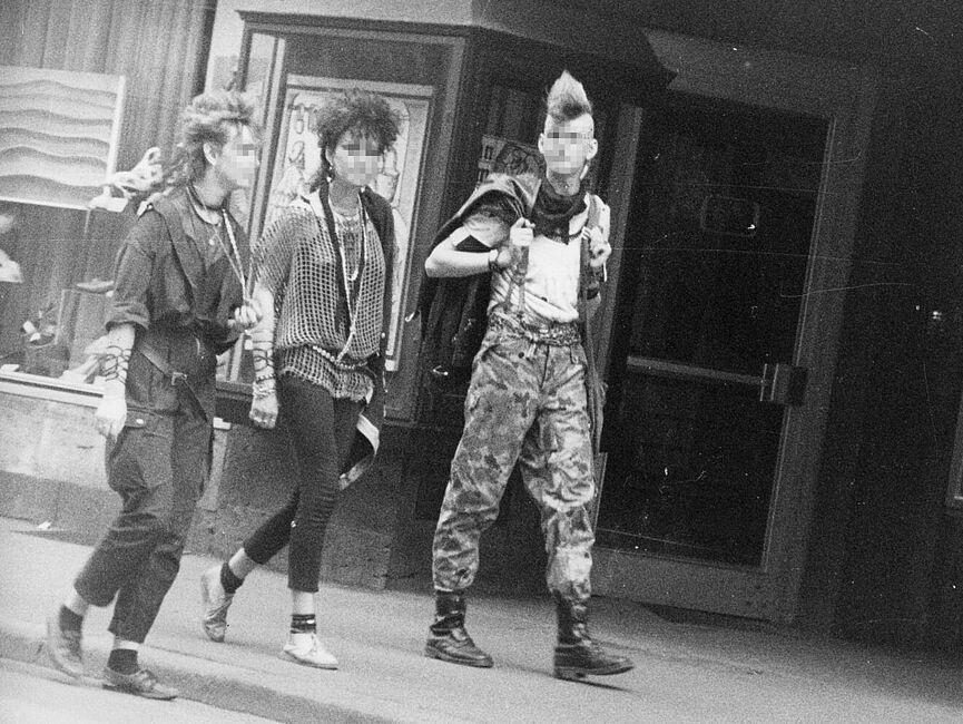 Schwarz-Weiß-Foto mehrerer Personen im Punk-Look auf einer Straße, die an der anscheinend versteckten Kamera vorbei laufen.