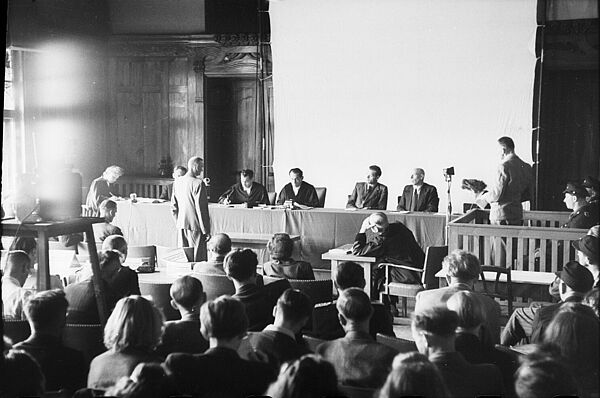 Auf dem Foto ist eine Reihe von Männern zu sehen, die nebeneinander an einem Tisch sitzt. Zwei der Männer tragen Talare. Vor ihnen steht ein Mann, der sie anschaut, Im Bildvordergrund sitzt eine Vielzahl an Menschen, die in Richtung der Männer schaut. Rechts am Bildrand steht ein Mann, vor ihm steht ein Mikrofon, hinter ihm sitzen zwei uniformierte Männer.