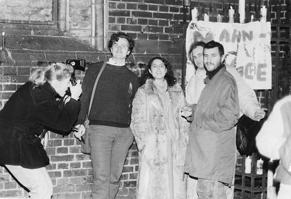 Eine Menschengruppe steht neben einer Kirche aus hellem Backstein während sie seitlich von einer Frau Fotografiert werden. Sie lächeln jedoch in die Kamera, mit der dieses Foto aufgenommen wurde.