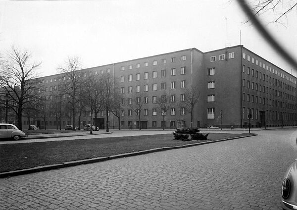 Blick über einen winterlichen Platz auf ein grau verputztes, sechsgeschossiges Gebäude Platz