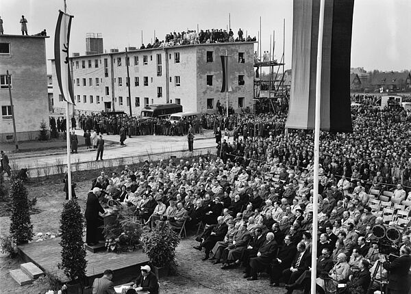 Einweihung des NAL Marienfelde am 14. April 1953; Quelle: picture-alliance / akg, Fotograf: Gert Schütz; Quelle: picture-alliance / akg, Fotograf: Gert Schütz Schwarz-weiß Foto: Vor den Rohbauten des Notaufnahmelagers sitzen die Gäste der Einweihung vor einem Redner an einem festlich geschmückten Rednerpult. Dahinter stehen weitere Zuschauer, auch auf dem Dach eines Gebäudes.