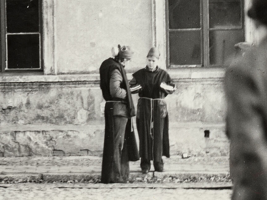 Pfarrer Freimark und Ehefrau mit Mönchskutten und Gasmasken vor der Pfarrei Schwarz-weiß Foto aus der Bilddokumentation der Stasi: Vor einer Hauswand mit zwei gr0ßen Fenstern stehen ein Mann und eine Frau, die Mönchskutten tragen und Gasmasken wie Hüte aufgesetzt haben.