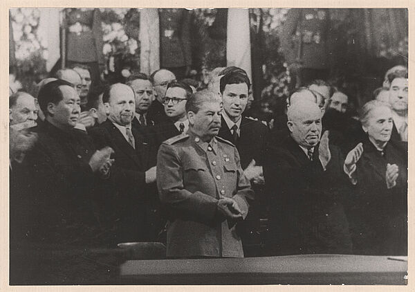 Schwarz-Weiß-Foto mehrerer anzugtragender Personen, die applaudieren. In der Bildmitte ist, in Uniform, Josef Stalin zu sehen.