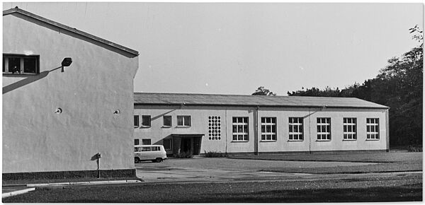 Gebäude des "Objekts 76" der Stasi im brandenburgischen Helenenau, um 1980 Blick über eine Rasenfläche auf ein rechtwinklig aus zwei Flügeln bestehndes, schmuckloses Gebäude mit zwei Geschossen. Die Stirnwand des linken Gebäudeflügels ist im Vordegrund zu sehen. Rechts daneben ist über eine gepflasterte Fläche der rechte Flügel zu sehen. Davor steht ein Kleinbus.