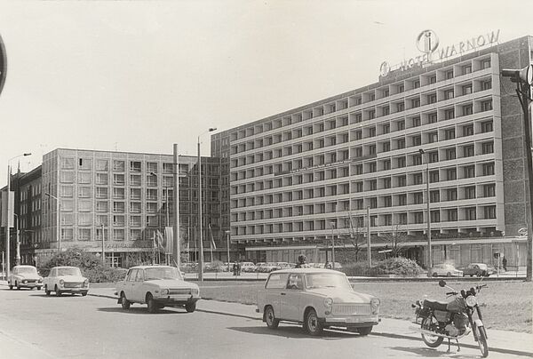 Schwarz-Weiß-Foto eines mehrstöckigen Plattenbaus. An der Fassade sind Balkons zu erkennen, auf dem Dach des Gebäude steht in großen Buchstaben "Hotel Warnow", auf einem Parkplatz stehen viele Autos. Im Vordergrund ist eine Straße zu sehen, an der mehrere Trabants, eine Simson u. Ä. parken.