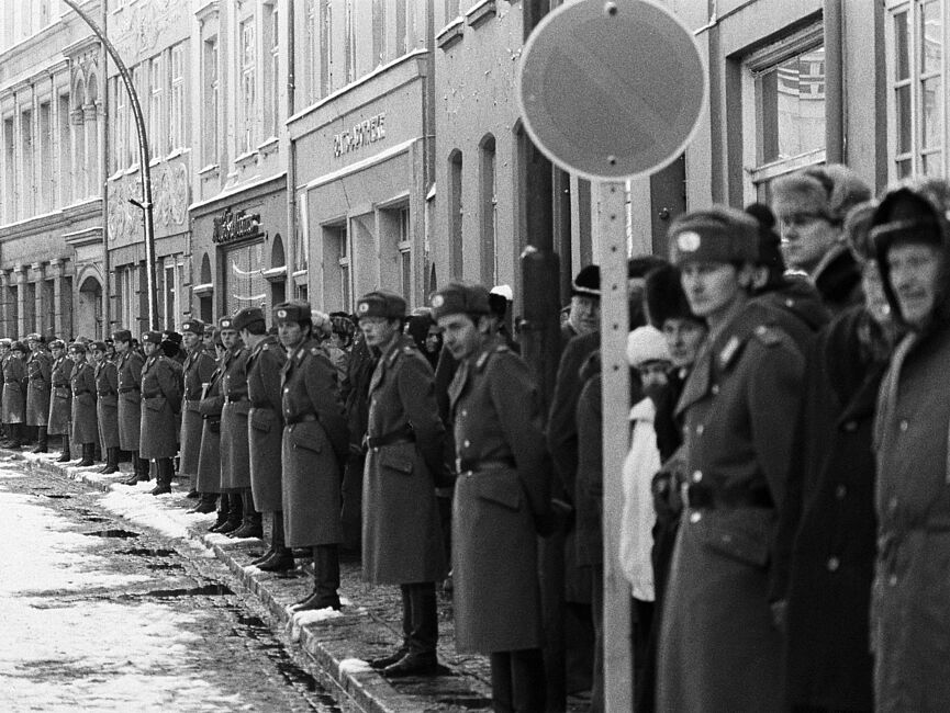 Blick eine nach links gekurvte Straße hinunter. Nur der rechte Straßenrand ist zu sehen, dahinter Gebäude. Am Straßenrand stehen Schulter an Schulter uniformierte Volkspolizisten Spalier, dahinter einige Schaulustige.