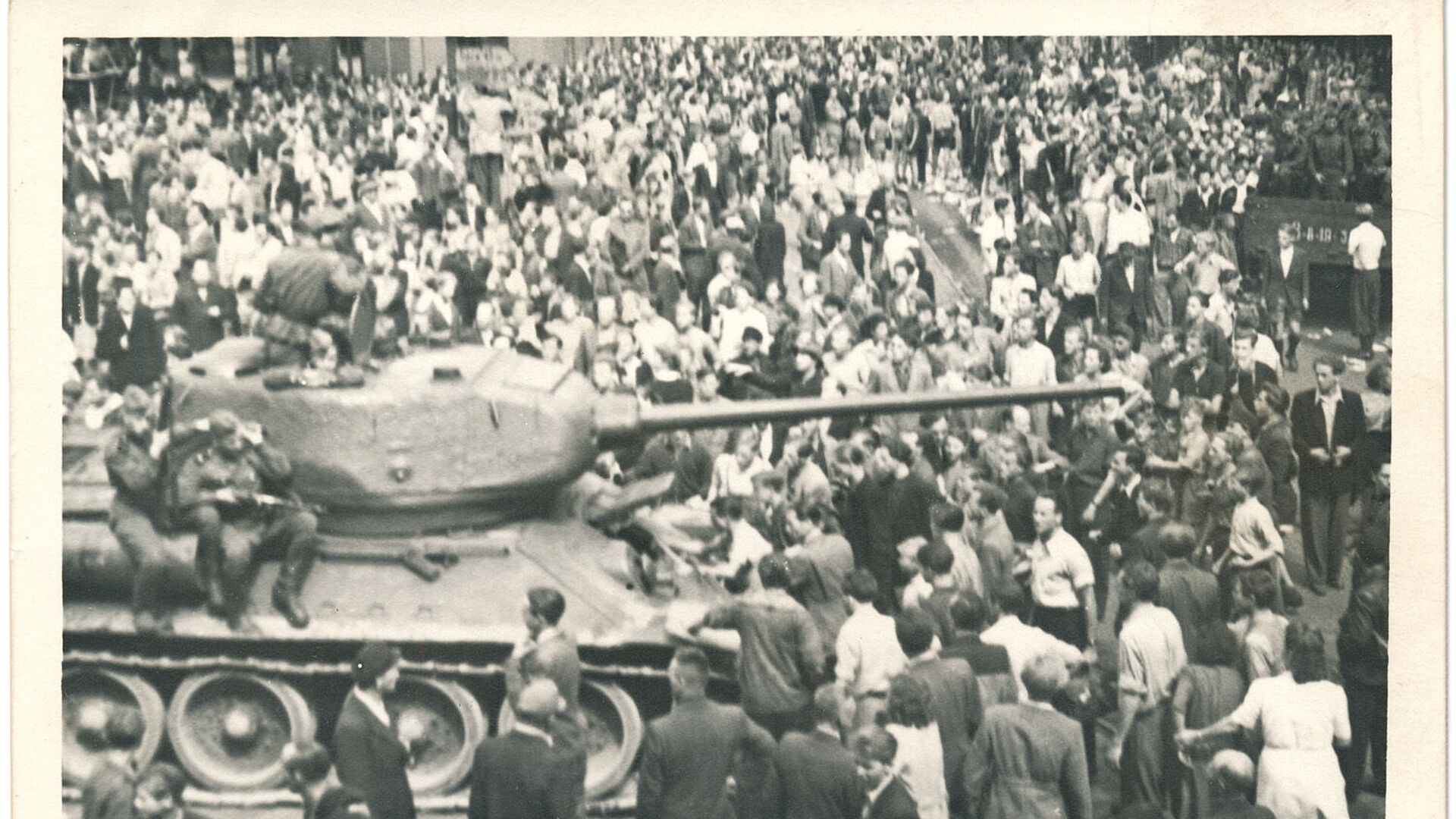 Schwarz-Weiß-Foto einer Menschenmenge, die einen Panzer umringt, auf dem bewaffnete Soldaten sitzen.