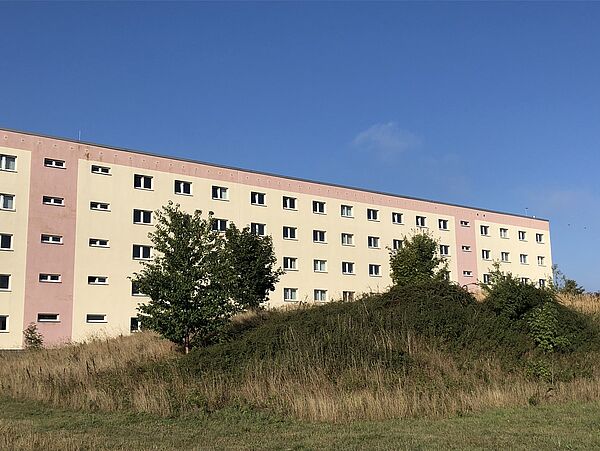 Farbfoto eines fünfstöckigen Flachdachbaus, der in den Farben Beige und Terrakotta gestrichen ist. Im Vordergrund sind eine Wiese und Hügel zu sehen, auf dem Gräse und Büsche wachsen. Der Himmel ist blau. 