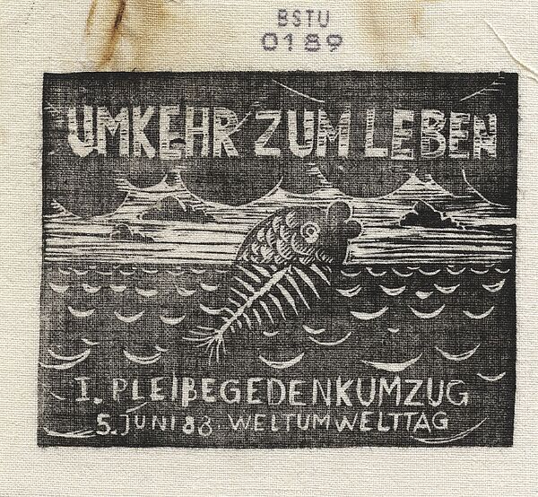 Bedrucktes Stoffstück, auf dem ein Fisch abgebildet ist, der aus dem Wasser herausschaut. Der untere, sich im Wasser befindliche Teil des Fisches besthet nur aus Gräten. Darüber steht der Text "Umkehr zum Leben", darunter "1. Pleißegedenkumzug, 5. Juni 