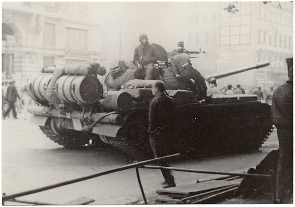 Schwarzweißbild eines Sowjetischen Panzersmit zwei Soldaten darauf. Im Hinergrund sieht man andeutungsweise eine Menschenmenger, die von links nach rechts geht. Im Vordergrund shteht ein halbabgewandter Mann.