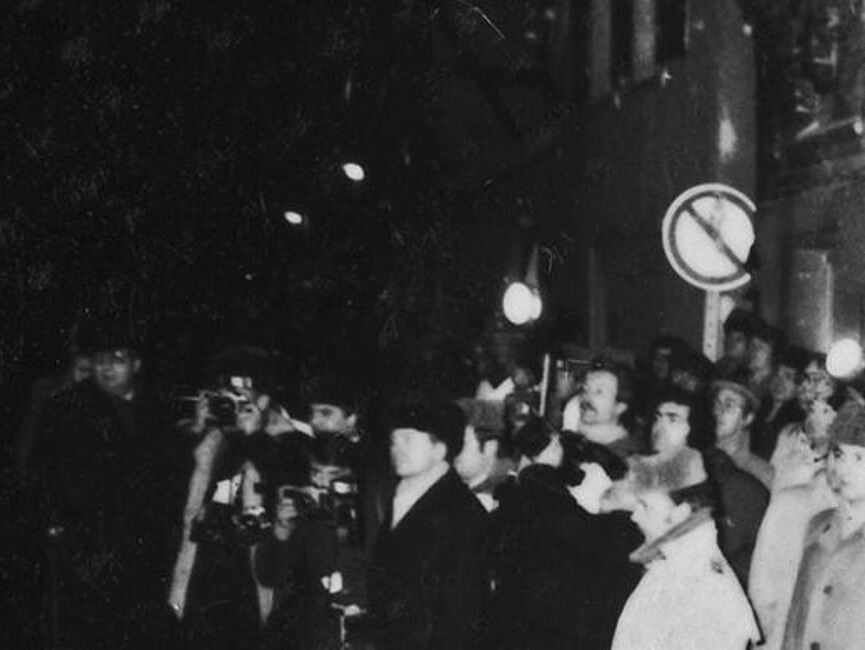 Ankunft der Wagenkolonne mit Bundeskanzler Schmidt und DDR-Staatschef Honecker am Weihnachtsmarkt in Güstrow: Schaulustige winken und halten Blumensträuße hoch, uniformierte Volkspolizisten überwachen die Menge, Pressefotografen machen Fotos. im Vordergrund ist das Heck eines Autos zu sehen