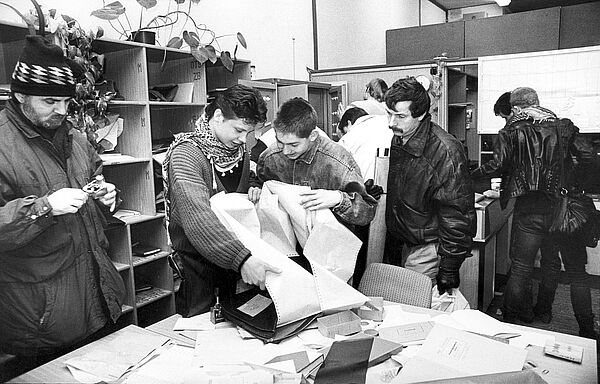 Mehrere Personen stehen in einem Büroraum. Zwei von ihnen halten Papier in der Händ, der Tisch vor ihnen ist voll mit weiteren Zetteln.