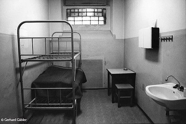 Zelle in der ehemaligen Untersuchungshaftanstalt der Staatssicherheit in Leipzig/ Beethovenstraße; Quelle: Gerhard Gäbler Schwarz-Weiß-Foto, blick in eine Zelle. An der Wand kommt durch ein paar Glasfliesen Licht in den Raum. Davor steht rechts ein kleiner Tisch mit Hocker, links ein Etagenbettgestell, auf der unteren Liege liegt eine Matratze. Im Vordergrund rechts ist ein Waschbecken.,