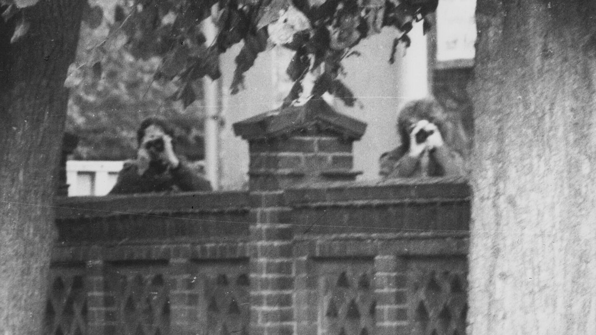 Schwarz-Weiß-Foto: zwei Männer schauen mit Ferngläsern über eine Mauer in Richtung des Fotografen.